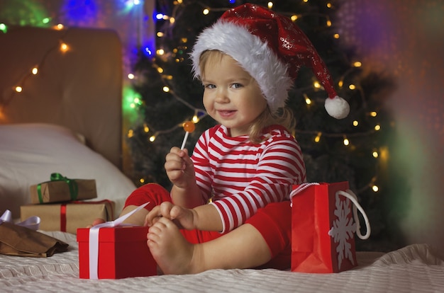 Kleines Mädchen in Weihnachtsmütze öffnet Weihnachtsgeschenkbox Lustiges und ausdrucksstarkes Kleinkind, das Geschenke genießt