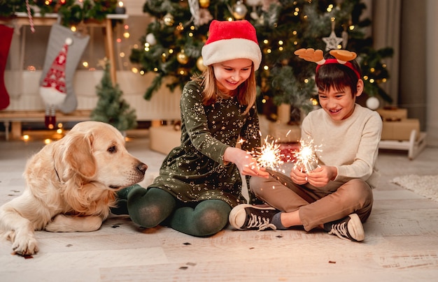 Kleines mädchen in weihnachtsmütze und junge mit rentierhörnern, die wunderkerzen halten, während sie zu hause neben golden retriever hund sitzen