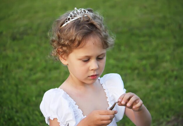 Kleines Mädchen in weißem Kleid mit Krone auf dem Kopf, das draußen auf dem grünen Sommerfeld auf dem Land spazieren geht