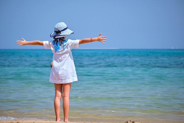 Kleines Mädchen in weißem Kleid und Hut, das barfuß mit weit gespreizten Händen am Strand steht und tropische Ferien mit Blick auf das Meerwasser genießt