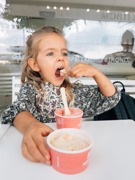 Kleines Mädchen isst Eis aus zwei Tassen mit einem Spatel am Tisch