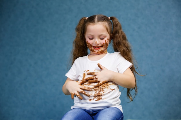 Kleines Mädchen isst Schokolade und verschmiert ihr weißes T-Shirt, Gesicht und reicht ihm die Hand