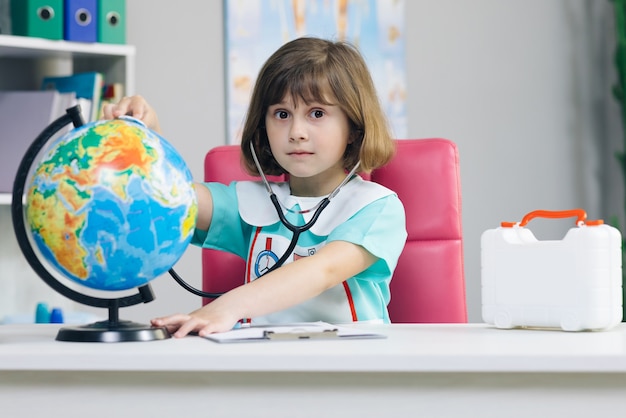 Kleines Mädchen ist in medizinischer Kleidung mit einem Stethoskop in den Händen gekleidet, untersucht und spielt ein Spiel, um den Planeten Erde zu heilen.