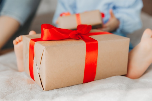 Kleines Mädchen Kleinkind sitzt auf dem Bett und zu ihren Füßen ist eine Schachtel mit einem Geschenk mit einem roten Band.