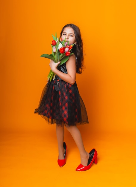 Kleines Mädchen lächelt in einem Kleid und großen roten Schuhen hält einen Blumenstrauß auf gelbem Hintergrund Platz für Text