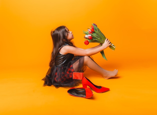 Kleines Mädchen lächelt in einem Kleid und großen roten Schuhen hält einen Blumenstrauß auf gelbem Hintergrund Platz für Text