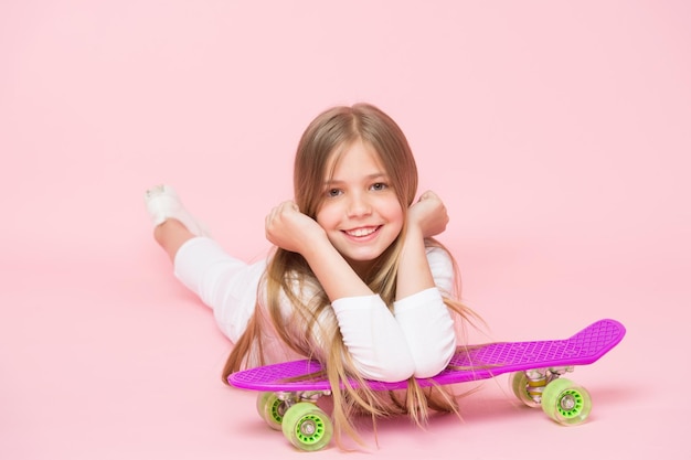 Kleines Mädchen lächelt mit Skateboard auf rosa Hintergrund Kinderskater lächelt mit Longboard Skateboard-Kind liegt auf dem Boden Kindheitslebensstil und aktive Spiele Sportliche Aktivität und energiegeladene Pastellfarben
