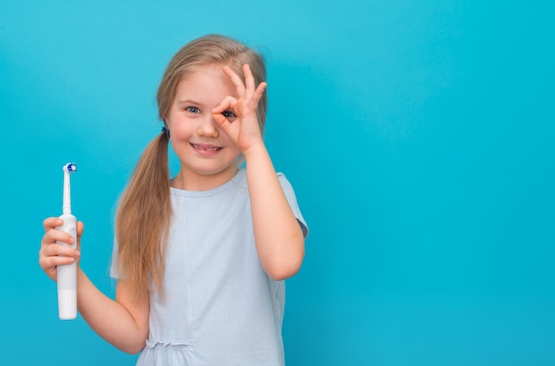 Kleines Mädchen lächelt und hält eine elektrische Bürste, die ein Ok-Zeichen auf blauem Hintergrund zeigt