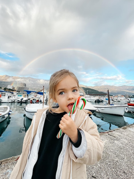 Kleines Mädchen leckt einen Lutscher auf einem Pier vor dem Hintergrund eines Regenbogens