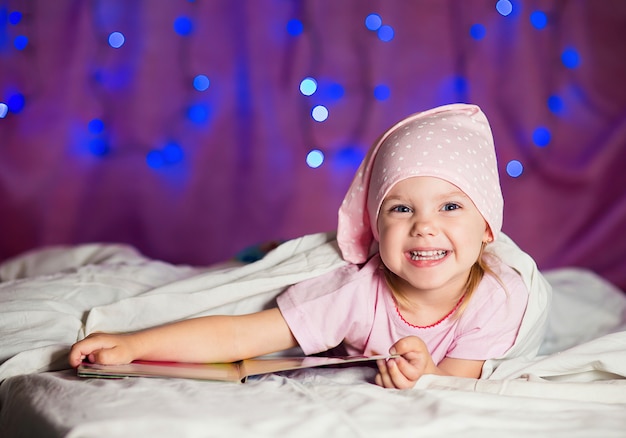 kleines Mädchen liegt im Bett in einem Schlummertrunk mit einem Kinderbuch und lächelt