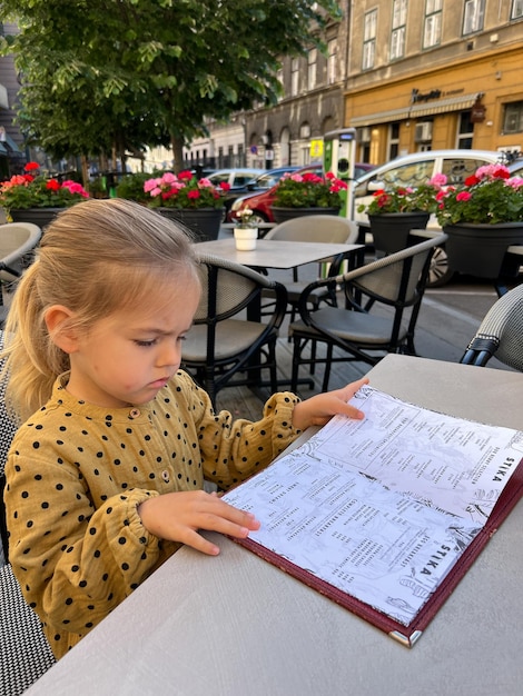 Kleines Mädchen liest eine Speisekarte an einem Tisch in einem Openair-Restaurant