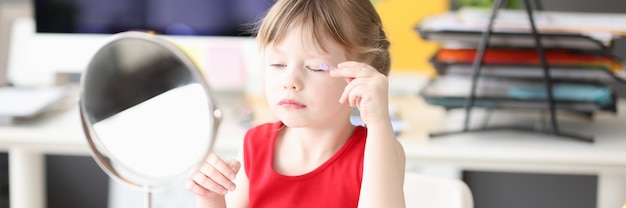 Kleines Mädchen malt ihre Augen und schaut im Spiegelbabykosmetikkonzept