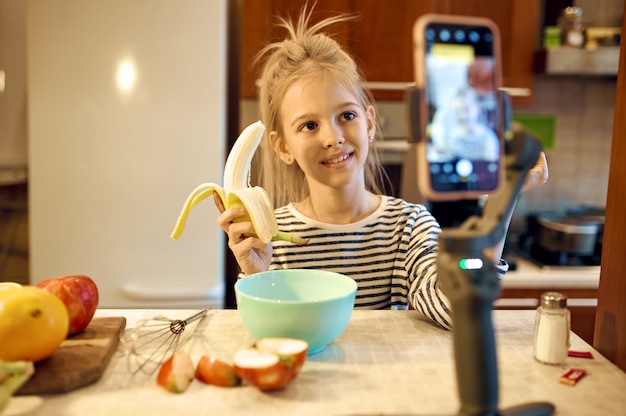 Kleines Mädchen mit Banane macht Lebensmittelblog, Kinderblogger. Kinderbloggen im Heimstudio, soziale Medien für junges Publikum, Online-Internetübertragung, kreatives Hobby