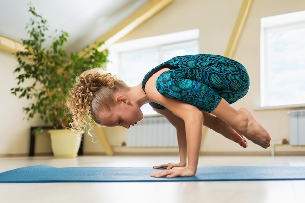 Kleines Mädchen mit blondem Haar, das Yoga praktiziert, das Handstandübung Kakasana oder Krähenhaltung auf einer Gymnastikmatte tut