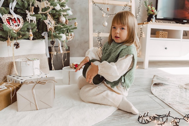 Kleines Mädchen mit blonden Haaren sitzt mit Geschenken auf dem Boden neben dem Weihnachtsbaum.