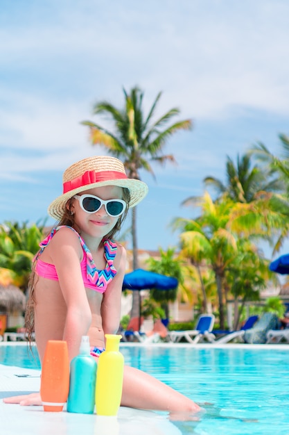 Kleines Mädchen mit der Flasche Sonnencreme sitzend am Rand des Swimmingpools