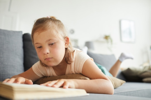 Kleines Mädchen mit Down-Syndrom, das auf Sofa liegt und ein Buch im Wohnzimmer liest