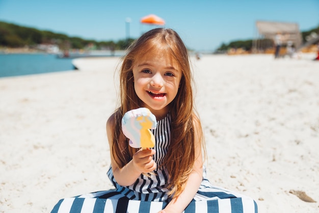 Kleines Mädchen mit Down-Syndrom in einem Sommerkleid isst Eis am Strand
