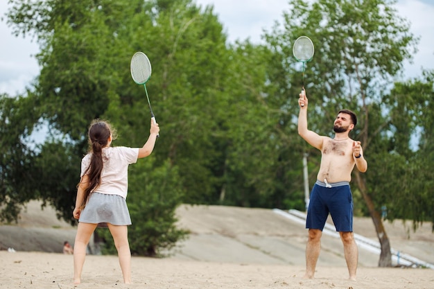 Kleines Mädchen mit dunklem Haar, das weißes T-Shirt trägt, grauer Rock, der Badminton spielt, mit einem bärtigen, hemdlosen Mann mittleren Alters