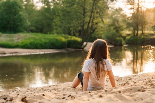 Foto kleines mädchen mit dunklen haaren sitzt auf dem sand in der nähe des waldes weibliches kind in weißem t-shirt und schwarzen shorts macht spaß in der natur