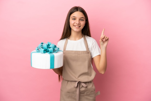 Kleines Mädchen mit einem großen Kuchen über isoliertem rosa Hintergrund, der einen Finger im Zeichen des Besten zeigt und hebt