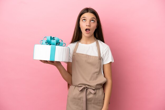 Kleines Mädchen mit einem großen Kuchen über isoliertem rosa Hintergrund, der nach oben schaut und mit überraschtem Ausdruck