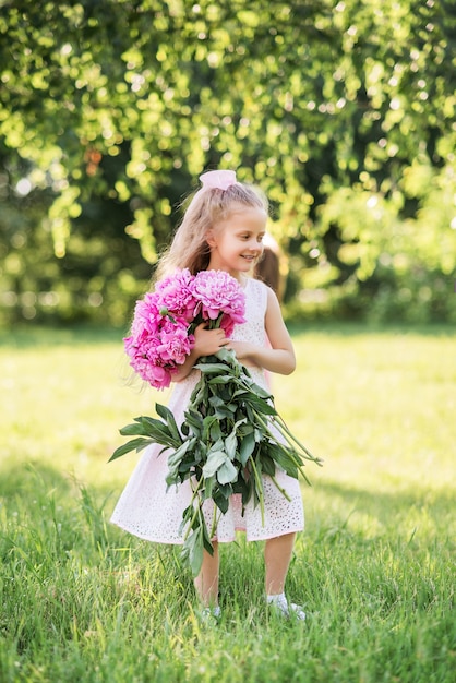 Kleines Mädchen mit einem großen Strauß Pfingstrosen. Ein Kind geht im Sommer mit Blumen im Park spazieren.