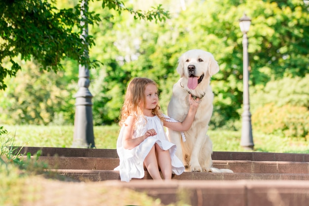 Kleines Mädchen mit einem großen weißen Hund im Park. Ein schönes 5-jähriges Mädchen im weißen Kleid umarmt ihren Lieblingshund während eines Sommerwegs.