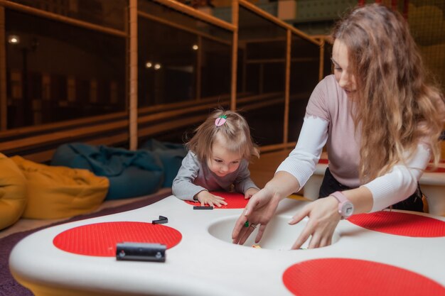 Kleines Mädchen mit einem Kindermädchen spielt Spielzeugsteine am Tisch in einem Kinderunterhaltungszentrum