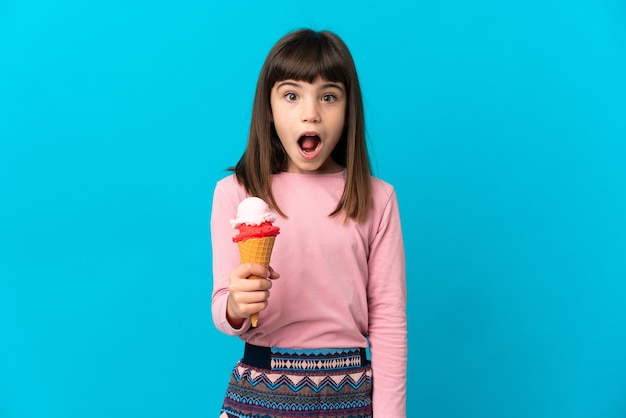 Kleines Mädchen mit einem Kornett-Eis isoliert auf blauem Hintergrund mit überraschendem Gesichtsausdruck