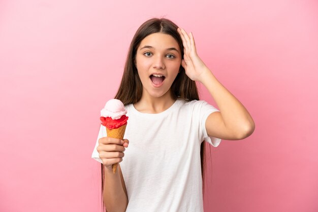 Kleines Mädchen mit einem Kornett-Eis über isoliertem rosa Hintergrund mit Überraschungsausdruck