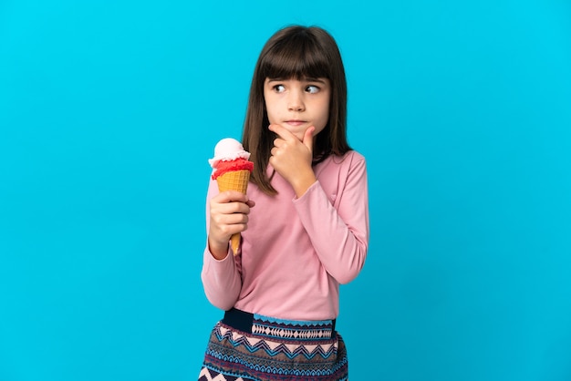 Kleines Mädchen mit einer Kornett-Eiscreme lokalisiert auf blauem Hintergrund, der Zweifel und Denken hat
