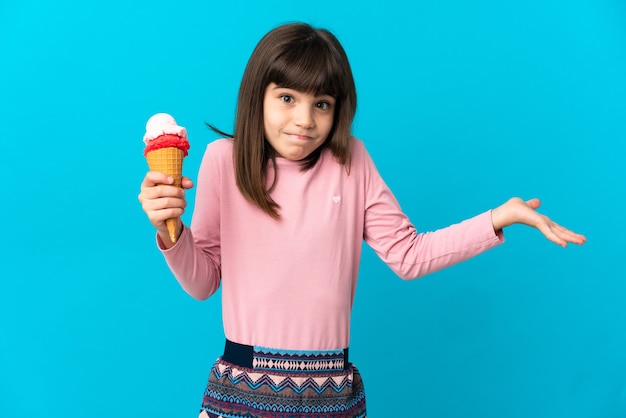 Kleines Mädchen mit einer Kornett-Eiscreme lokalisiert auf blauer Wand, die Zweifel beim Erhöhen der Hände hat