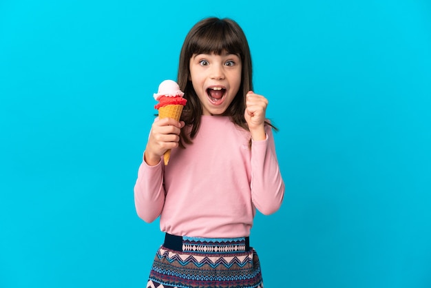 Kleines Mädchen mit einer Kornetteiscreme lokalisiert auf blauem Hintergrund, der einen Sieg in der Siegerposition feiert