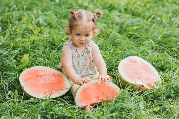 Kleines Mädchen mit einer lustigen Frisur isst eine Wassermelone auf dem Rasen im Park gesunder Snack für Kinder