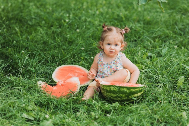 Kleines Mädchen mit einer lustigen Frisur isst eine Wassermelone auf dem Rasen im Park gesunder Snack für Kinder