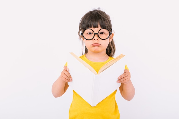 Kleines Mädchen mit ernster Geste mit gelbem T-Shirt und runder schwarzer Brille, die ein offenes weißes Buch in den Händen auf weißem Hintergrund hält