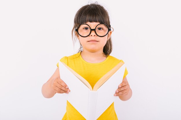 Kleines Mädchen mit gelbem T-Shirt und runder schwarzer Brille, das ein offenes Buch in den Händen auf weißem Hintergrund hält
