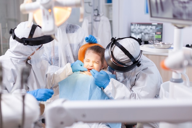 Kleines Mädchen mit geschlossenen Augen wegen Schmerzen während der Hohlraumbehandlung. Stomatologie-Team, das während der Covid19-Prozedur an Kinderzähnen einen PPE-Anzug trägt.