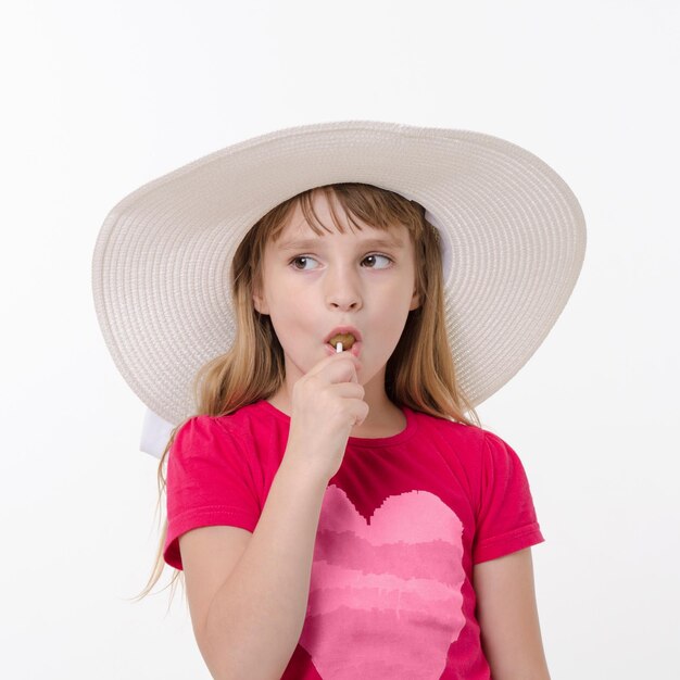Kleines Mädchen mit großem Hut, das Lutscher isst Studioportrait auf weißem Hintergrund