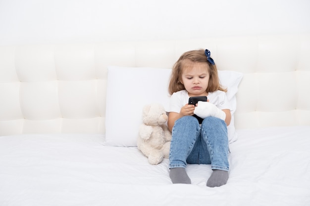 Kleines Mädchen mit Hand in der Besetzung, die im Bett unter Verwendung des Smartphones sitzt und Cartoon oder Bildungsvideo ansieht.