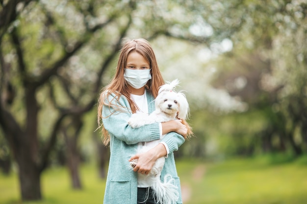 Kleines Mädchen mit Hund mit medizinischer Schutzmaske zur Vorbeugung von Viren im Freien im Park