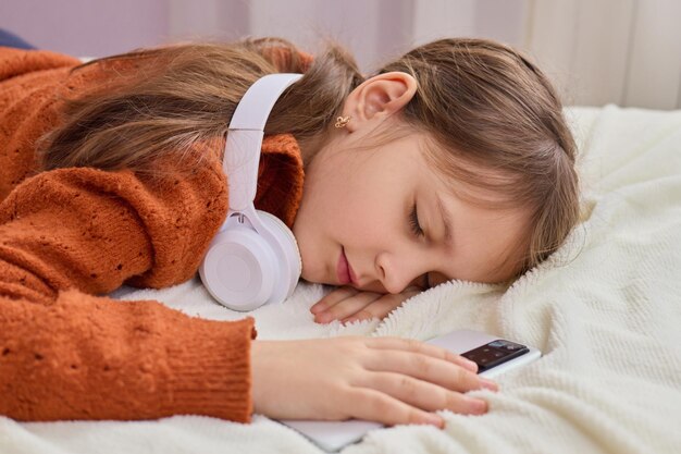 Foto kleines mädchen mit kopfhörern und smartphone, das lange stunden mit dem gerät schläft in ihrem schlafzimmer auf dem bett, liegt mit geschlossenen augen und genießt die tagesschläfrigkeit