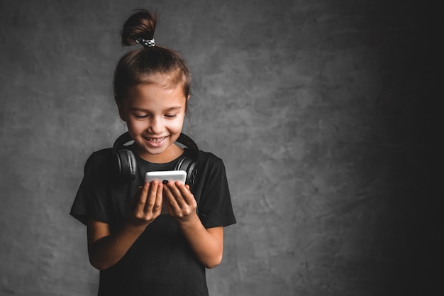 Kleines Mädchen mit Kopfhörern und Telefon auf grauem Hintergrund