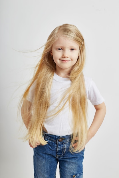 Kleines Mädchen mit langen blonden Haaren und in Jeans, die auf einer weißen Wand aufwerfen. Freude Spaß, junge Modell Kinder Mode. Modellschule für Kinder.