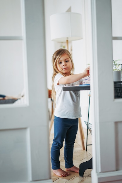 Kleines Mädchen mit langen Haaren in der Hauskleidung, die am Tisch steht und spielt, Wirkung des Guckens von Türen