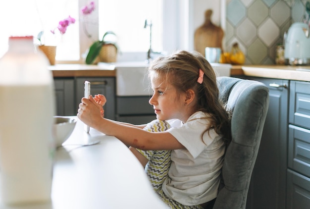 Kleines Mädchen mit langen Haaren in Hauskleidung mit Handy am Esstisch in der Küche zu Hause