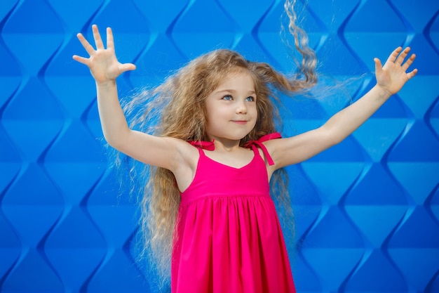 Kleines Mädchen mit langen lockigen Haaren in einem rosa Kleid auf blauem Hintergrund tanzt und lacht, helle Kindergefühle der Freude, glückliche Kindheit