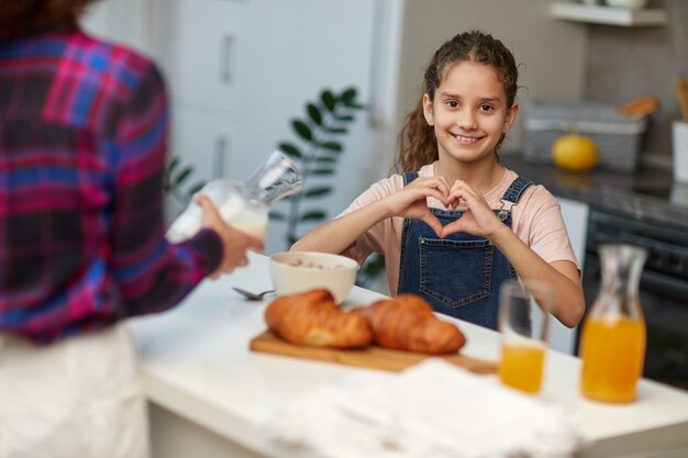 Kleines mädchen mit lockigem haar in der küche mit ihrer mutter, das die kamera anschaut und dabei herzsymbolform mit den händen lächelt, nimmt frühstück zusammen.