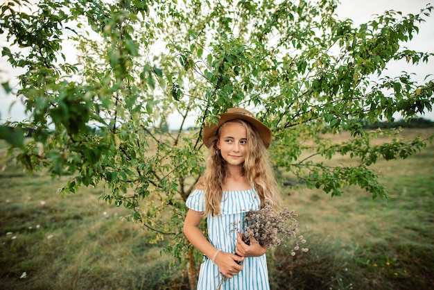 Kleines Mädchen mit lockigem Haar in einem Hut und Blumen steht in einem langen Kleid